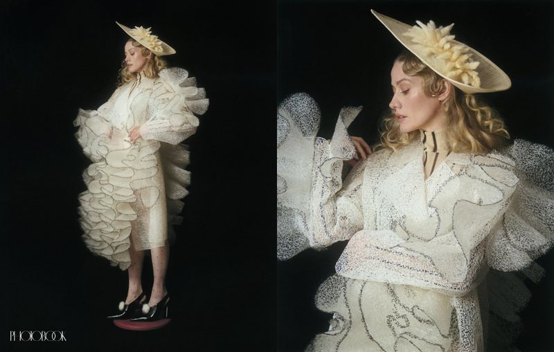 Olivia Taylor Dudley in a sculptural ruffled off-white gown and saucer hat against a black backdrop for Photobook Magazine.