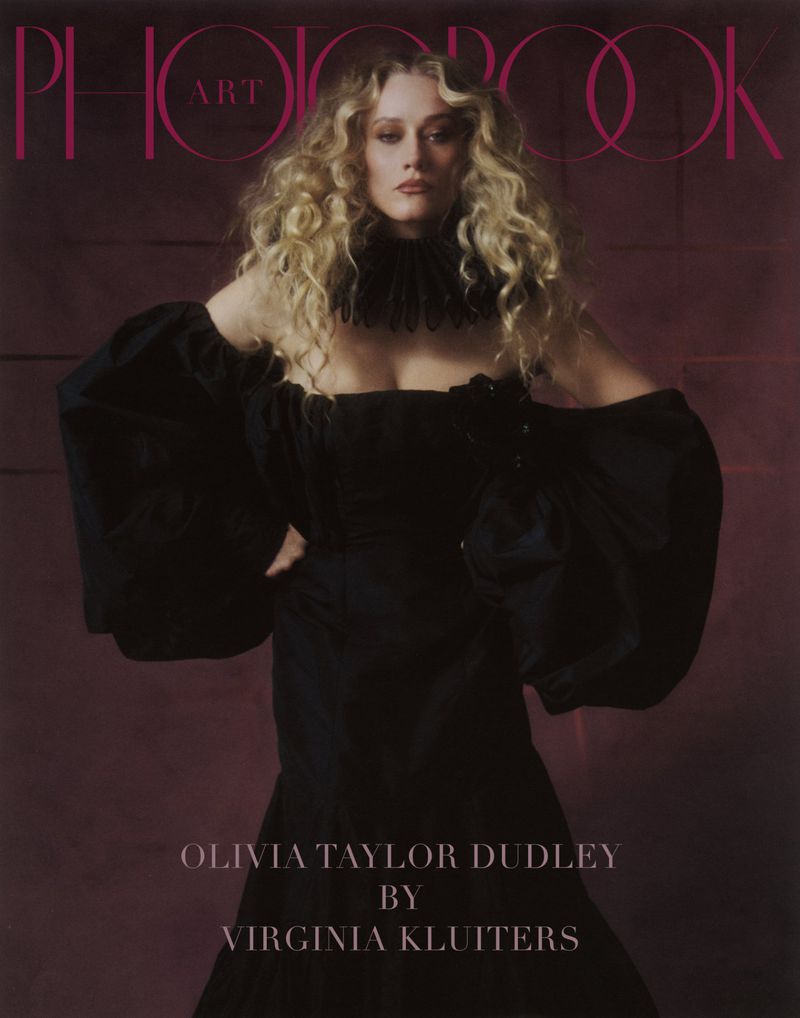 Olivia Taylor Dudley posing in a black off-shoulder gown with voluminous sleeves and a ruffled neckpiece for Photobook Magazine.