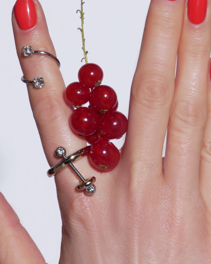 Close-up of a hand with red nail polish wearing jewelry rings, adorned with a sprig of redcurrants against a white backdrop.