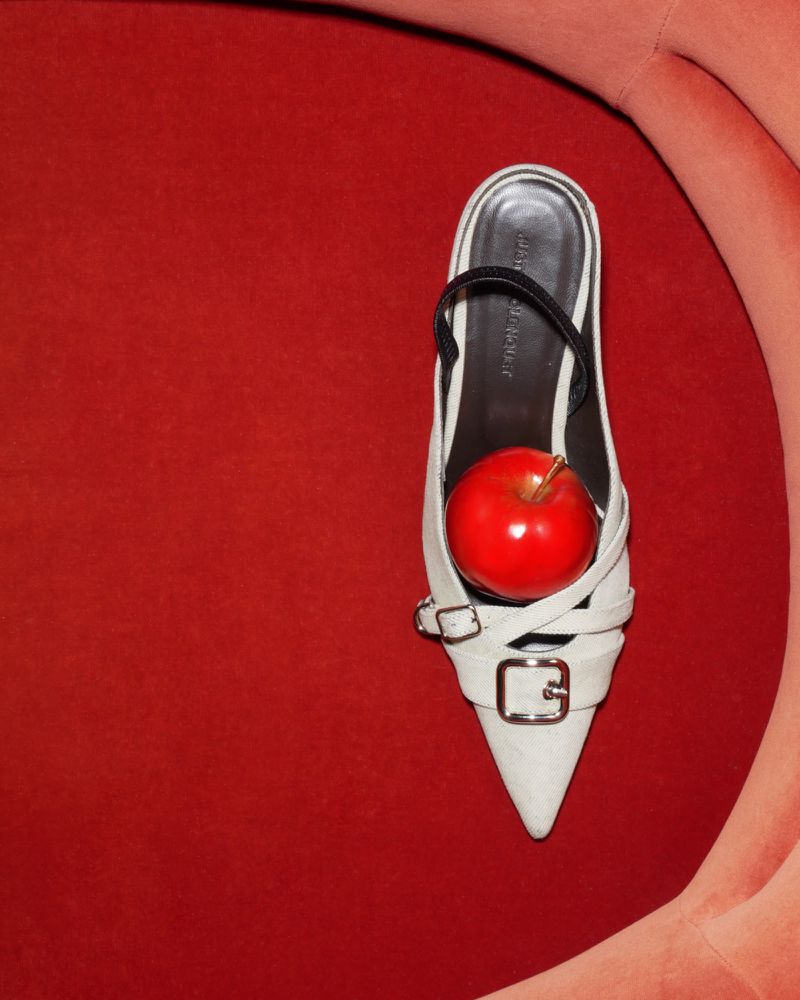 Elevated close-up of a white, pointed-toe high-heeled mule with a red apple balanced inside the shoe, against a red fabric background.