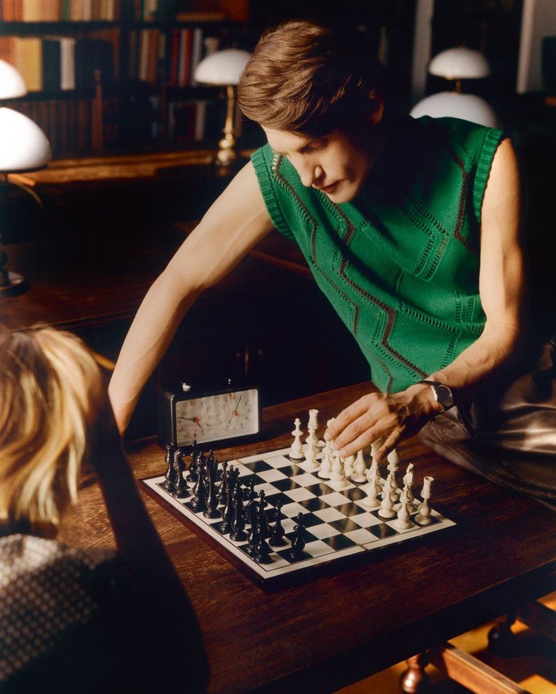 Model Anatol Modzelewski in a green sleeveless knit leaning over a chessboard opposite an opponent in a library for Vogue Polska.