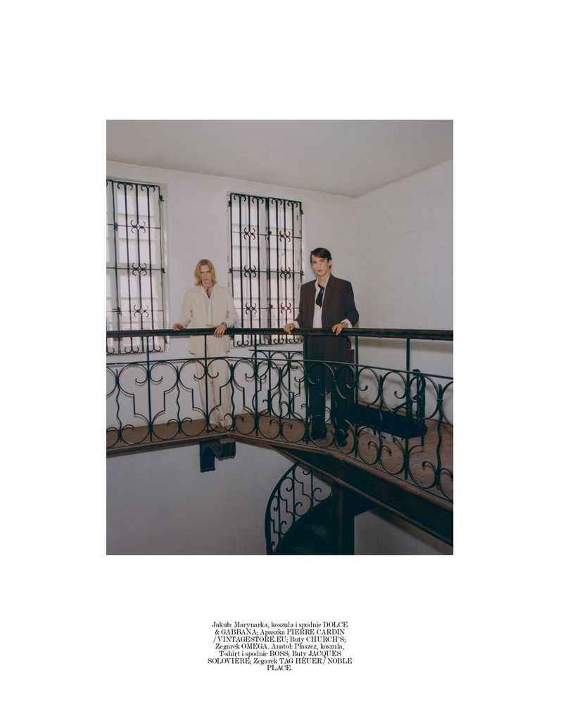 Two male models standing along a wrought-iron balcony railing above an interior spiral staircase.