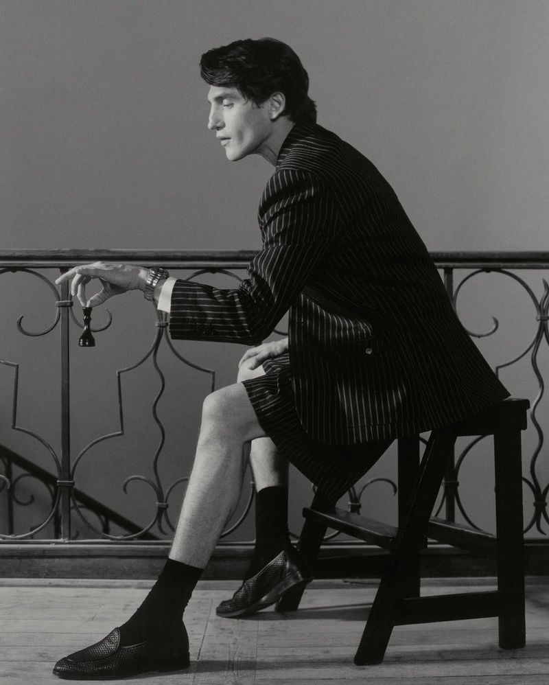 Anatol Modzelewski sits in a pinstripe blazer and shorts holding a chess piece beside an ornate iron railing.