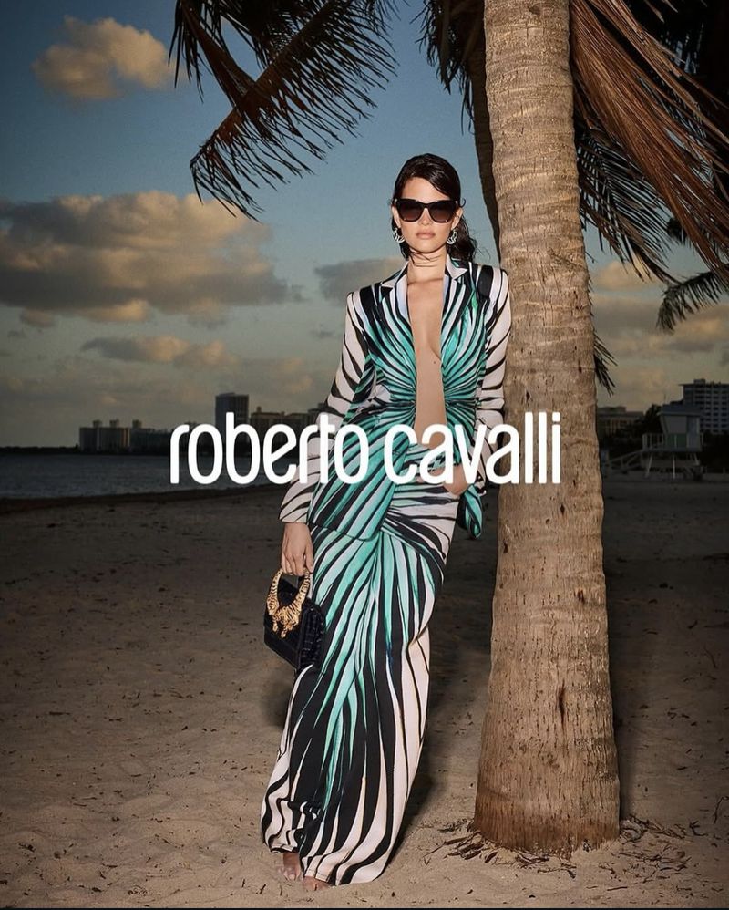 A stylish adult woman stands on a sandy beach next to a palm tree, wearing a patterned Roberto Cavalli dress, sunglasses, and holding a purse.