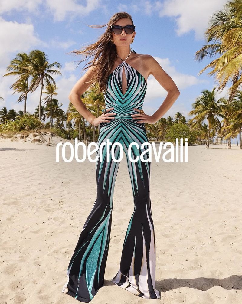A tall fashion model poses on a beach in a turquoise, black, and white patterned halter jumpsuit, sunglasses, and a necklace on a sunny day.