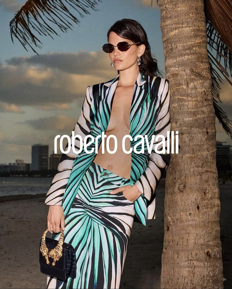 Woman in a palm-leaf-patterned suit poses on the beach next to a palm tree, holding a croc-skin purse with gold tiger heads.