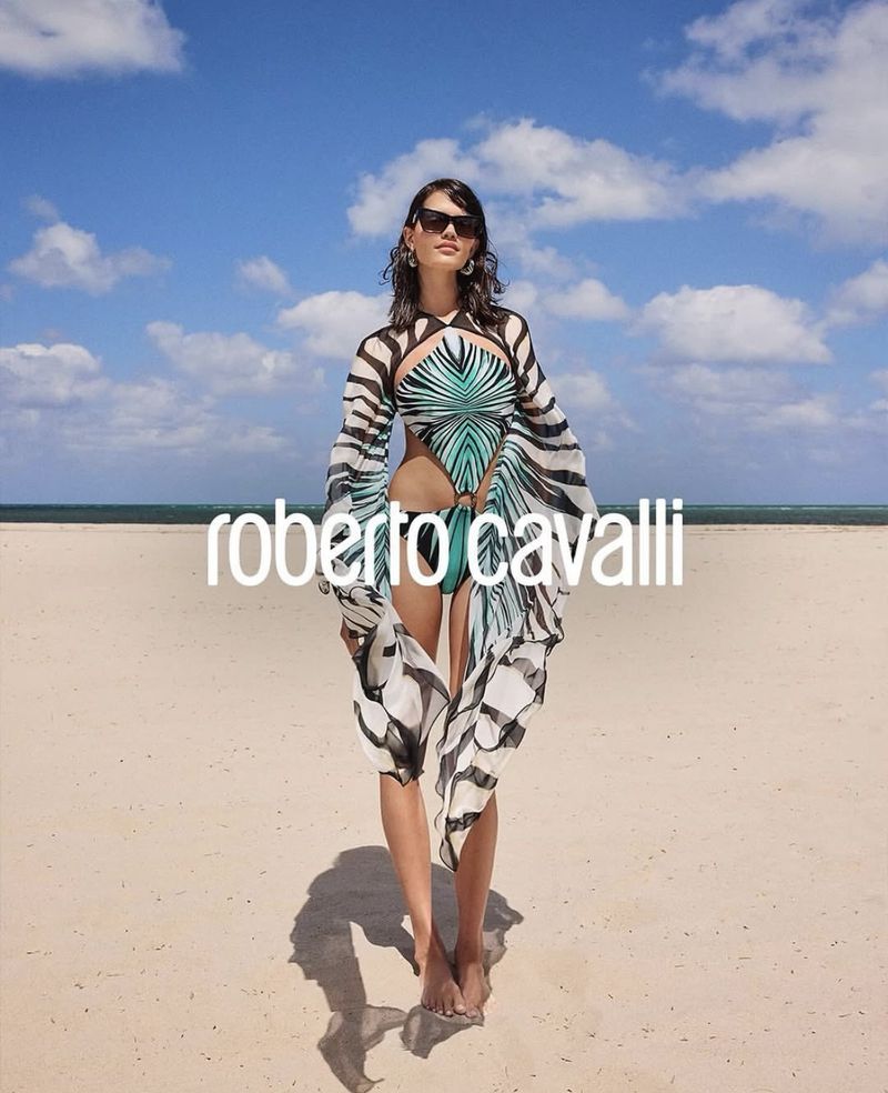 Woman in patterned swimsuit and cover-up poses on a sandy beach with a cloudy sky and turquoise ocean backdrop; fashion editorial shoot.