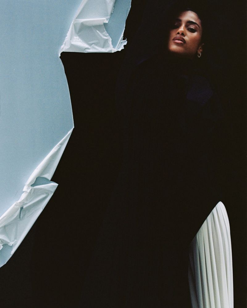 Model Imaan Hammam posing against a torn light blue paper backdrop wearing black fabric with white pleated trousers.
