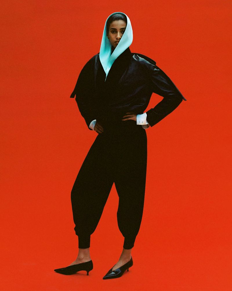 Imaan Hammam poses standing in a black outfit with a light structured hood against a solid red backdrop for Dazed MENA.