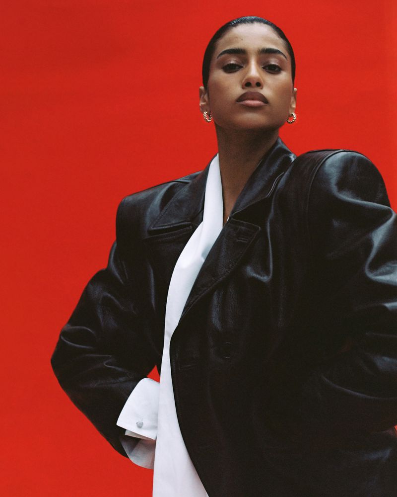 Model Imaan Hammam wearing an oversized black leather coat over a white collared shirt against a bold red background.