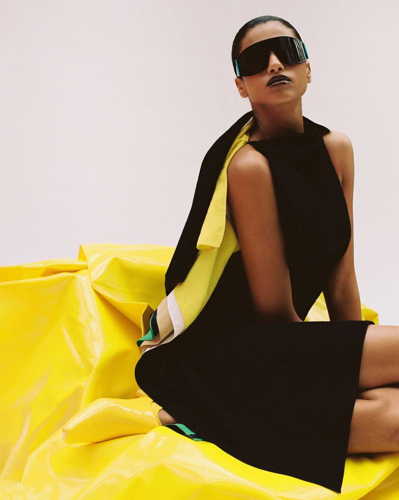 Imaan Hammam wearing a black dress and oversized shield visor sunglasses seated on glossy yellow vinyl for Dazed MENA.