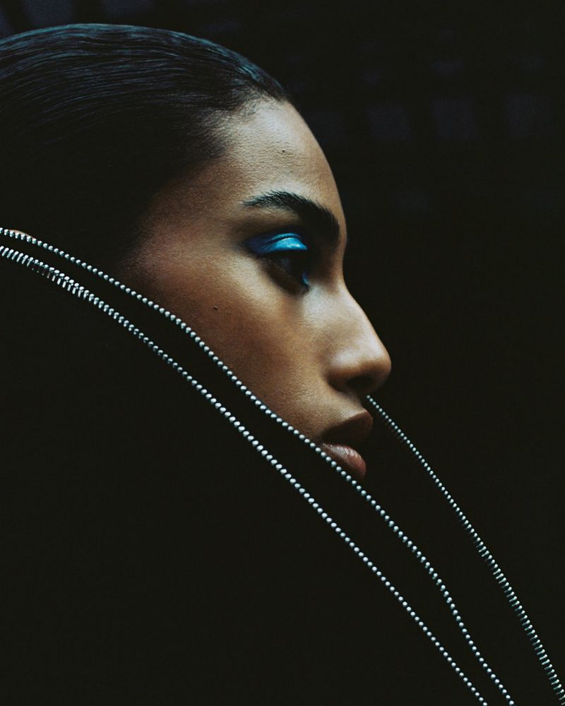 Close-up profile of model Imaan Hammam wearing vivid blue eyeshadow and a high black collar accented with metallic zippers.