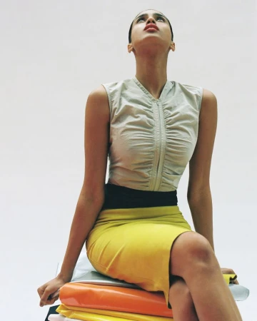 Model Imaan Hammam seated in a light grey ruched zip-up top and yellow skirt looking upwards against a white backdrop.