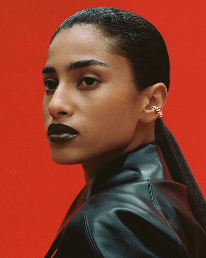 Imaan Hammam in profile wearing a black leather jacket and gold ear cuff against a vibrant red background.