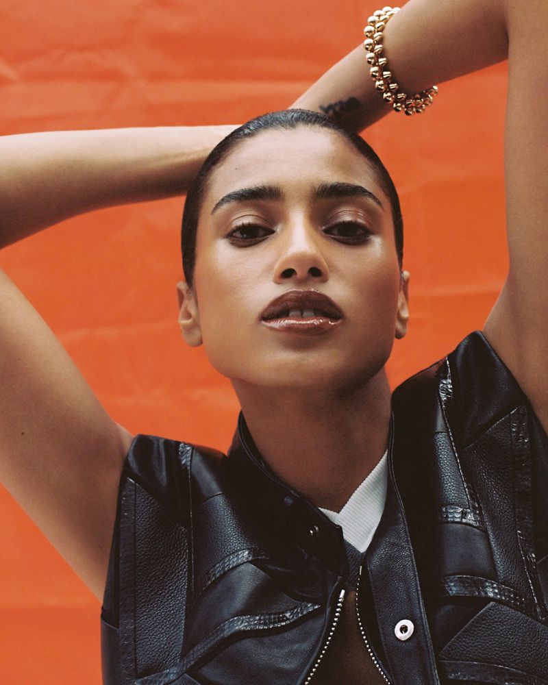 Model Imaan Hammam poses with hands behind head wearing a black leather vest and gold beaded bracelet against an orange backdrop.