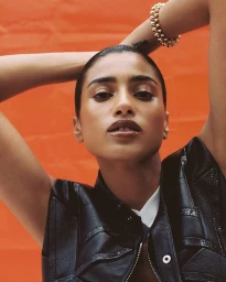 Model Imaan Hammam poses with hands behind head wearing a black leather vest and gold beaded bracelet against an orange backdrop.