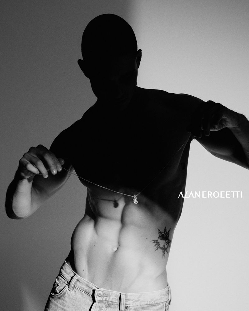 Black and white studio shot of a male model with a shadowed face, visible abs, wearing jeans and a pendant necklace.