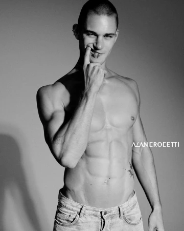 Black and white portrait of a shirtless adult male in light wash jeans with hand raised near his face against a gradient gray background.