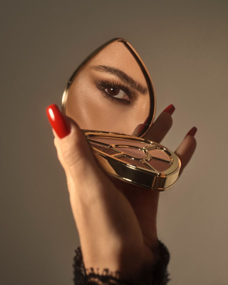 Hand with red manicured nails holding an open gold compact mirror reflecting a woman's smokey eye makeup against a warm studio background.
