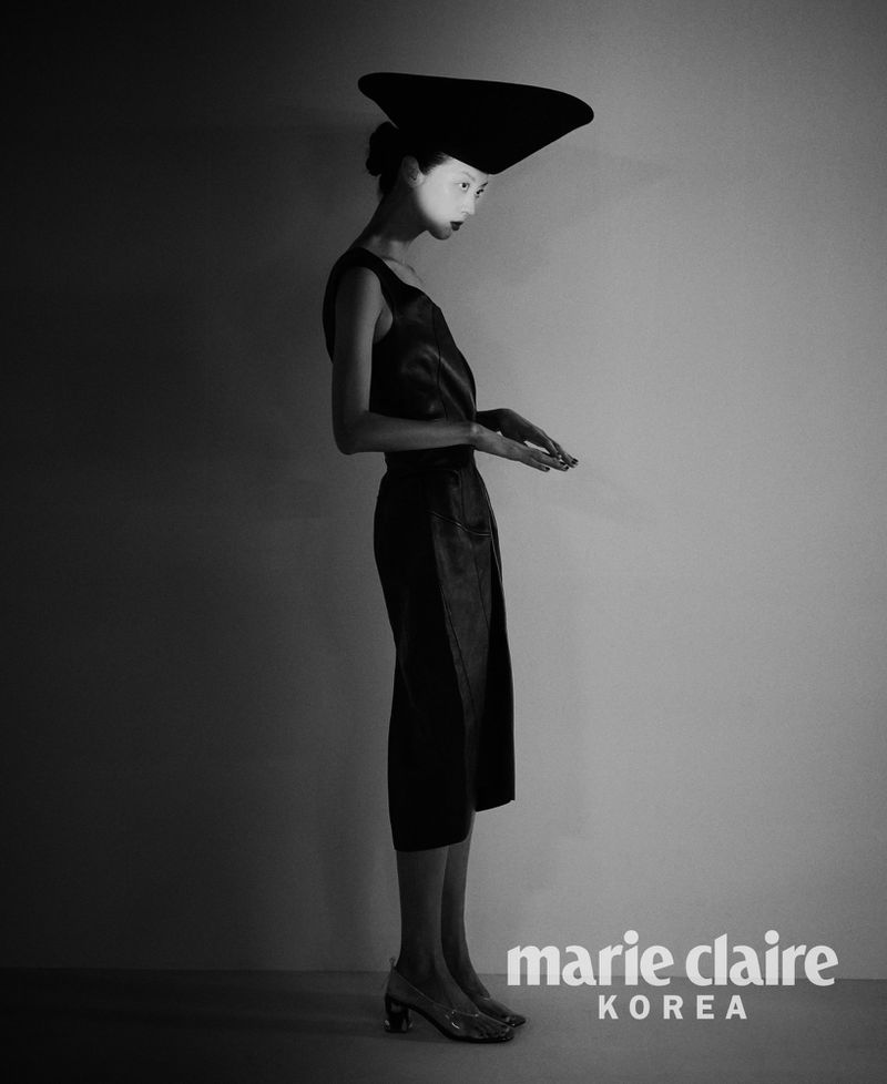Model So Young Kang posing in profile wearing an oversized sculptural black hat and sleeveless midi dress in black and white.