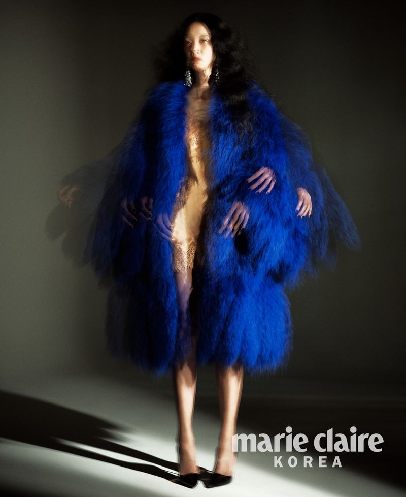 Model So Young Kang wearing a vibrant blue faux fur coat with multiple motion-blurred hands against a grey studio backdrop.