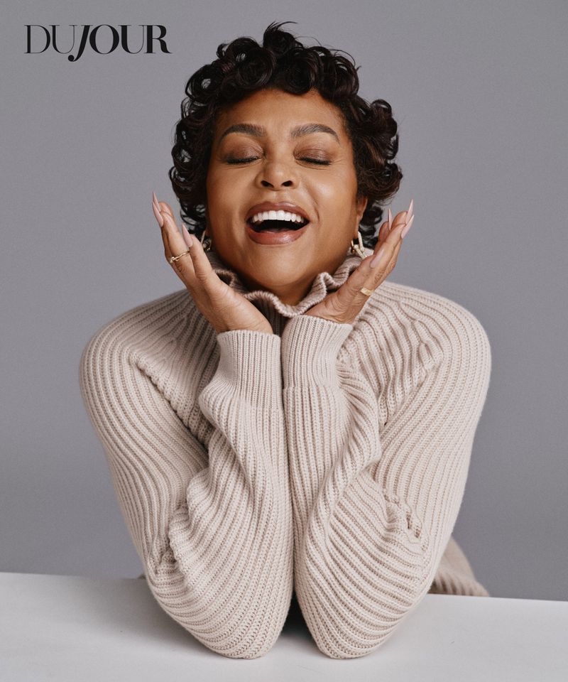 Taraji P. Henson laughing with hands framing her face while wearing a beige ribbed knit turtleneck against a grey backdrop.