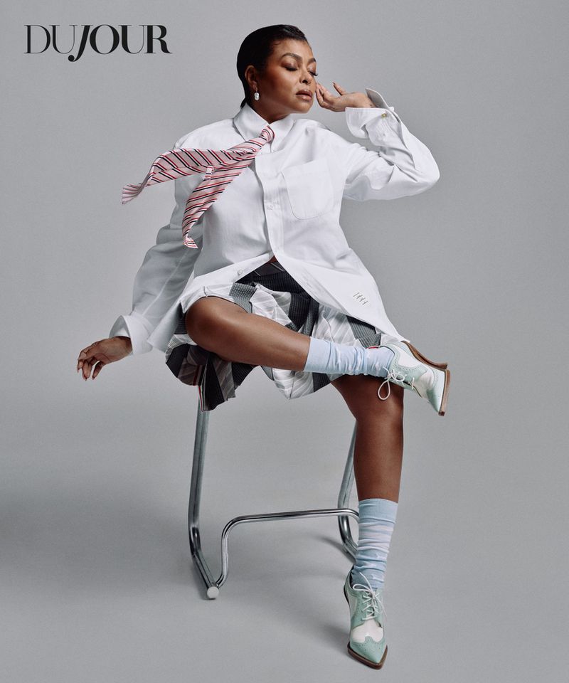 Taraji P. Henson seated on a metal chair wearing a white shirt, plaid shorts, striped tie, and socks against a grey background.
