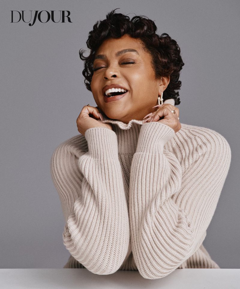 Taraji P. Henson laughing while holding the collar of a cream ribbed knit turtleneck sweater against a grey backdrop.