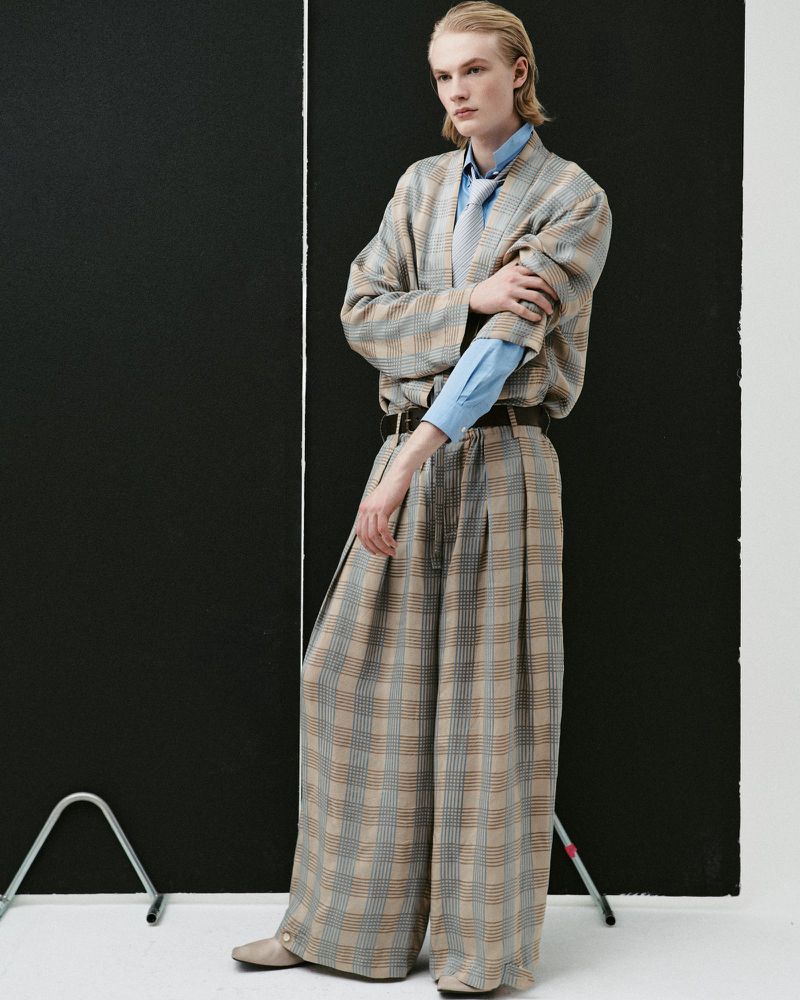 Model Leander Straub poses in an oversized plaid suit and blue shirt in front of black studio panels on a white floor.