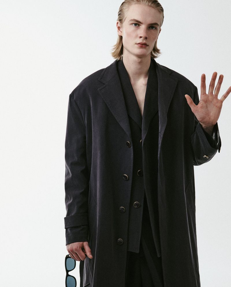 Model Leander Straub in an oversized dark overcoat raising his hand against a white seamless studio backdrop.