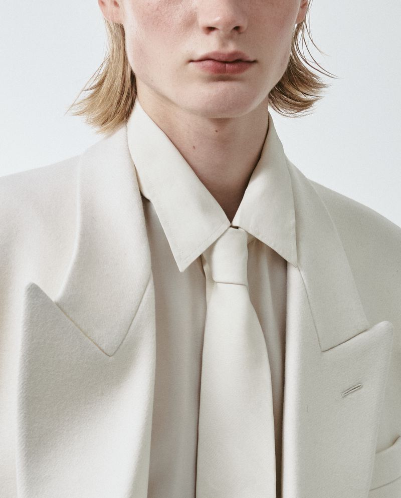 Close-up of a model wearing an off-white tailored suit jacket, collared shirt, and matching tie against a white background.