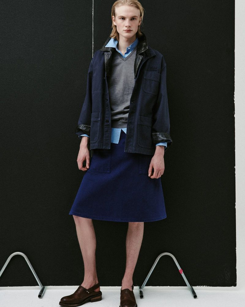 Model Leander Straub wearing a navy jacket, grey sweater, and blue knee-length skirt against a black panel background.