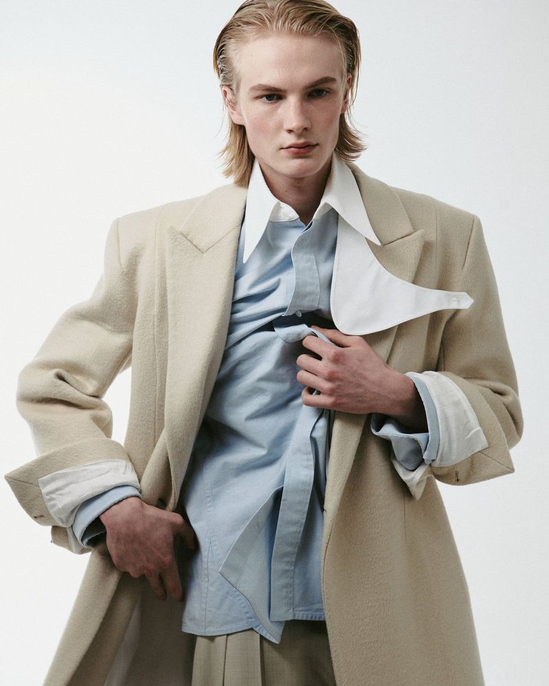 Model Leander Straub wearing an oversized beige wool coat with layered collared shirts against a white seamless background.