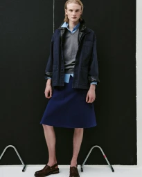 Model Leander Straub wearing a navy jacket, grey sweater, and blue knee-length skirt against a black panel background.