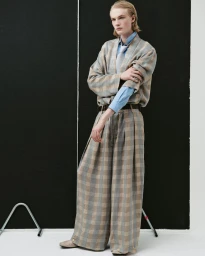 Model Leander Straub poses in an oversized plaid suit and blue shirt in front of black studio panels on a white floor.