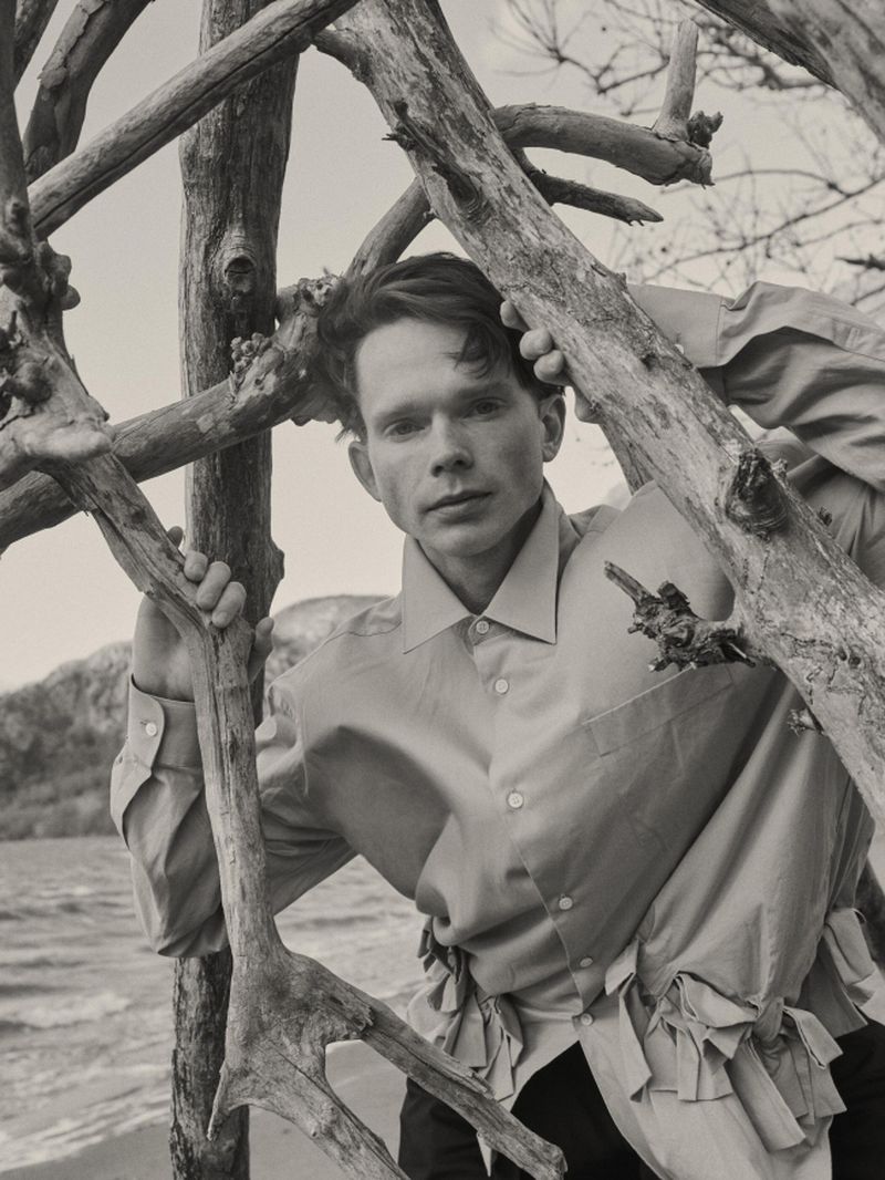 A man in a collared shirt posing behind weathered bare branches on a coastal shoreline.