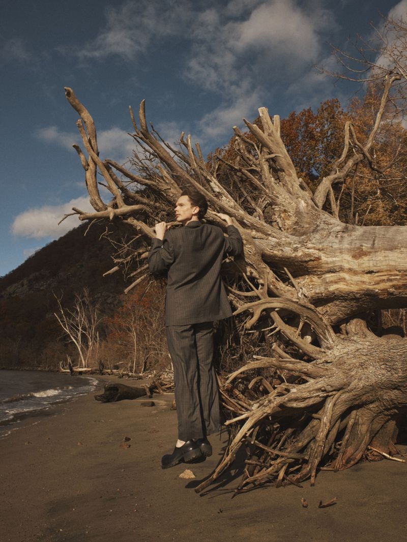 Model in a dark grey tailored suit standing beside a large fallen tree with exposed roots on a sandy shoreline.