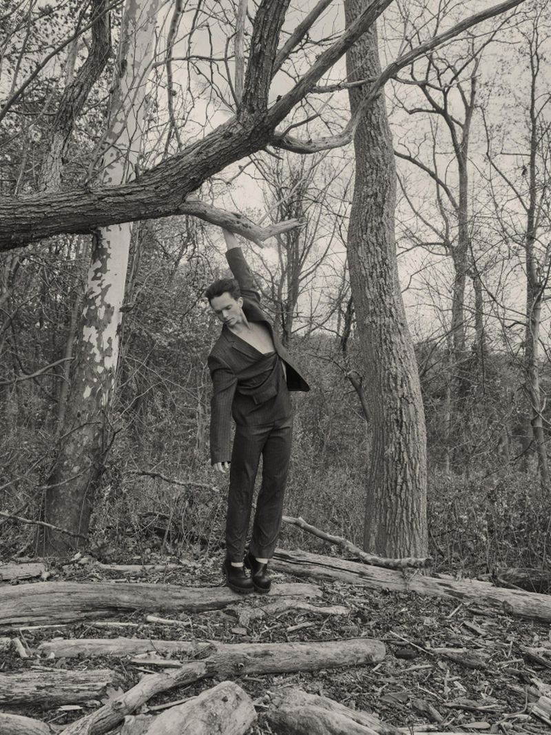 A man in a dark tailored suit poses while reaching up toward an overhead tree branch in a dense woodland setting.