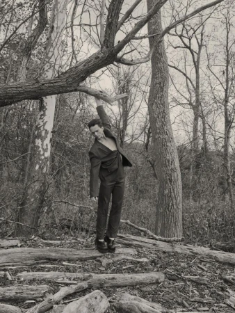 A man in a dark tailored suit poses while reaching up toward an overhead tree branch in a dense woodland setting.