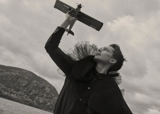 Low angle view of a young man wearing an avant-garde black ruffled outfit holding a toy biplane against an overcast sky.