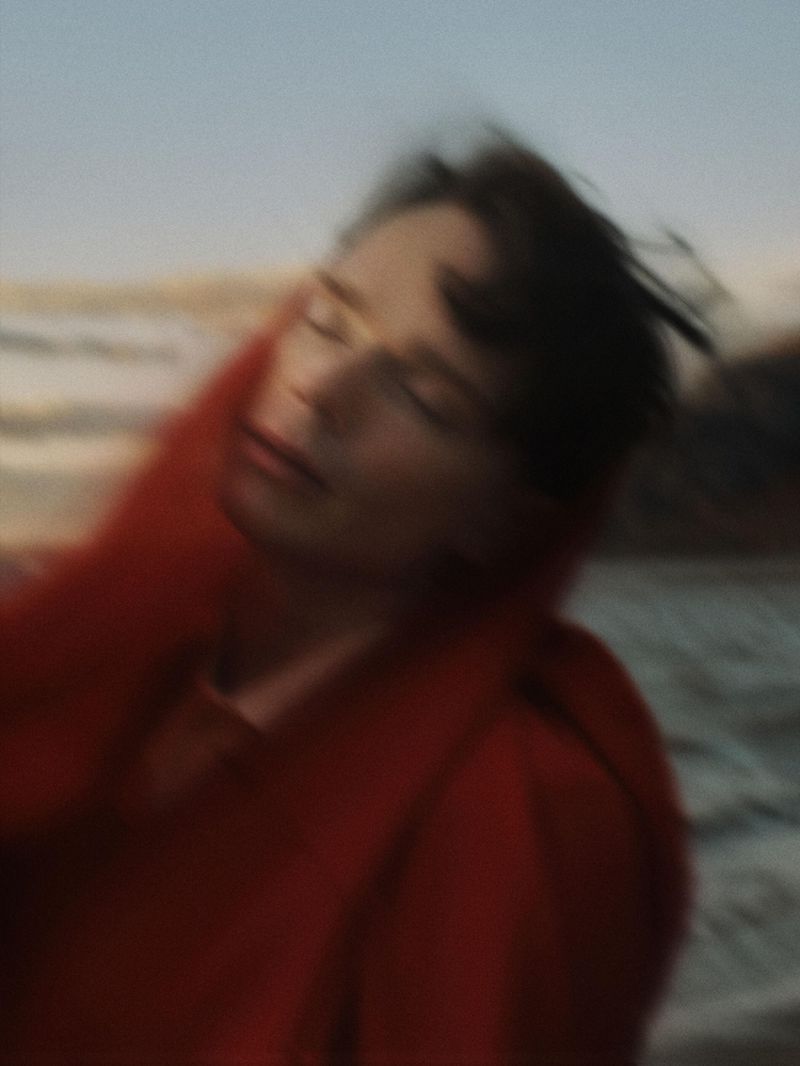 Motion-blurred portrait of a person with eyes closed draped in a vibrant red fabric along an outdoor coastal beach.