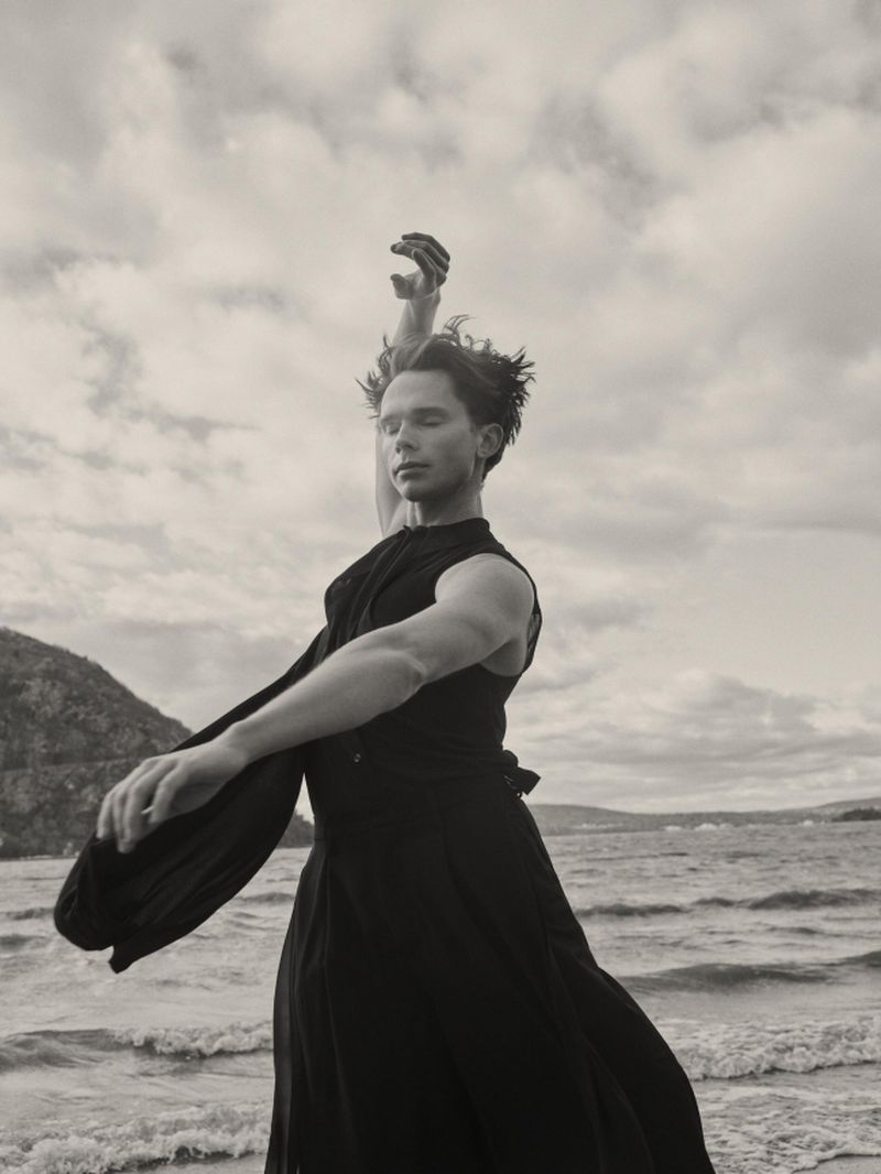 A man in a flowing black sleeveless garment poses with arms outstretched on a beach with water and hills behind him.