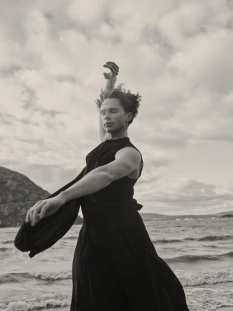 A man in a flowing black sleeveless garment poses with arms outstretched on a beach with water and hills behind him.