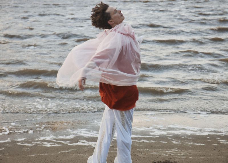 A person standing on a sandy shoreline wearing sheer billowing fabric and white pants with head tilted back toward the sky.
