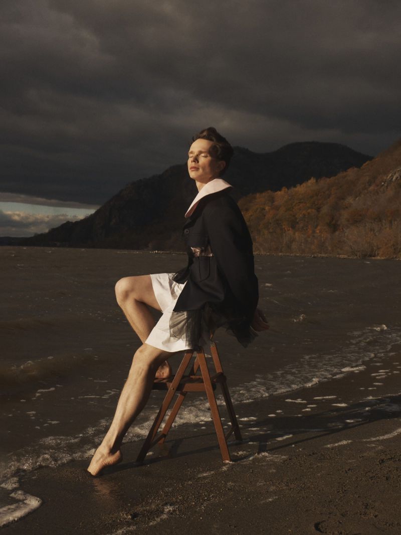 Model seated on a wooden chair at a sandy water shoreline wearing a black coat with a wide collar and white shorts in dramatic sunlight.