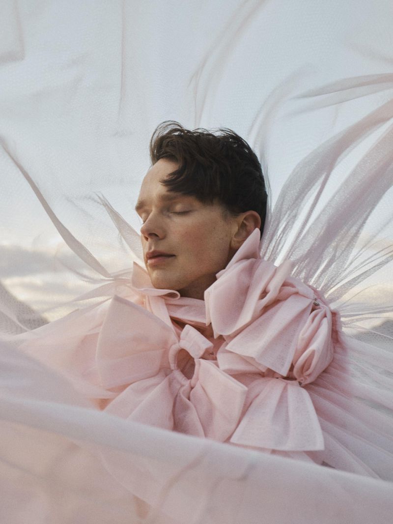 Close-up portrait of a man with closed eyes wearing a sculptural pastel pink ruffled collar enveloped in sheer billowing fabric.