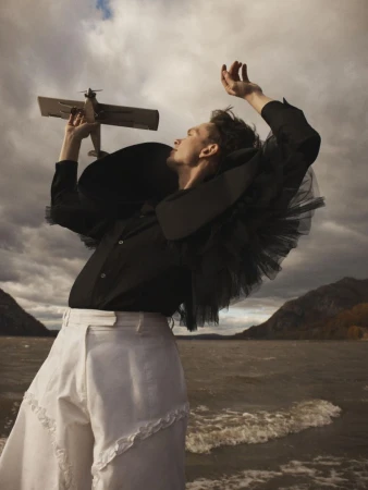 A man holding a toy airplane aloft by a lake, wearing a dramatic black ruffled tulle top and white trousers against an overcast sky.