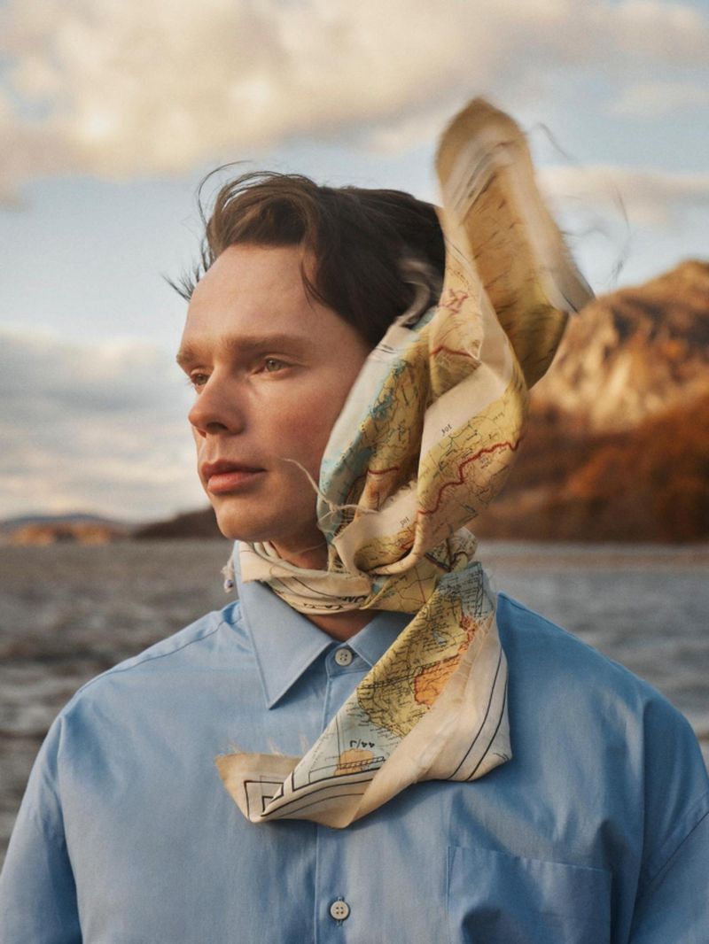 Man in a light blue buttoned shirt with a map printed silk scarf blowing in the wind around his head against a lake and mountain backdrop.