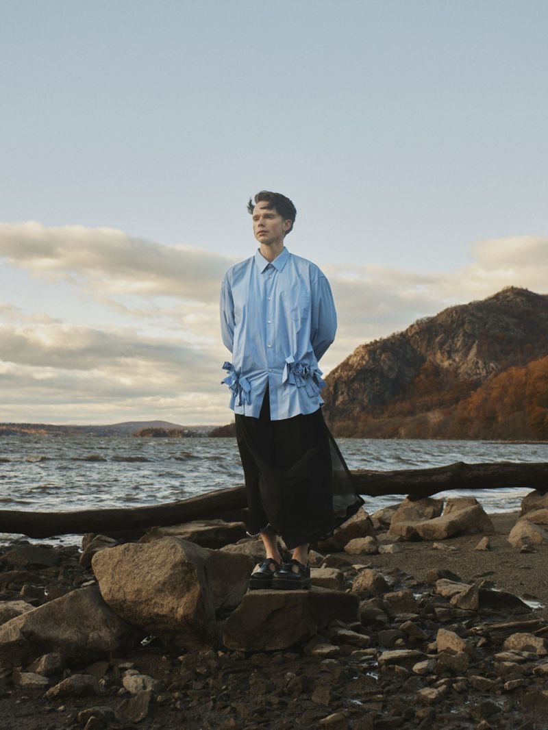 A young man wearing a light blue tied shirt and black sheer skirt stands on a rocky riverbank in front of distant hills.