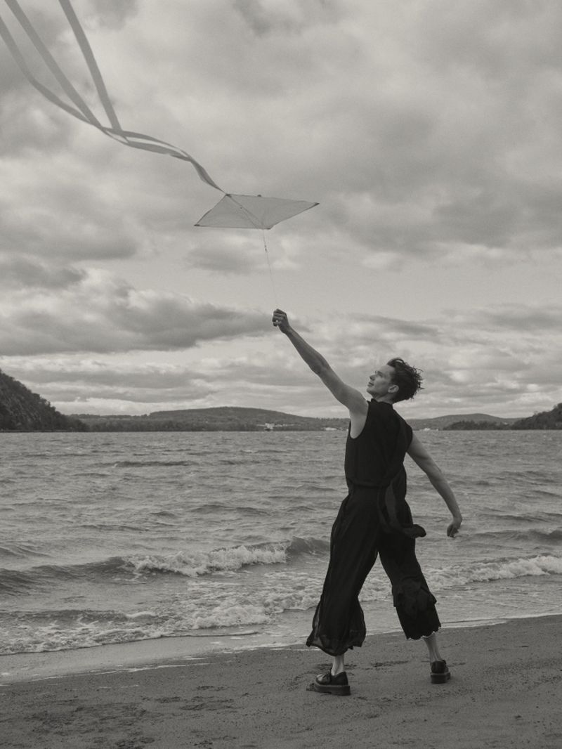 Person in a black sleeveless outfit flying a white kite with long tails along a windy lakeshore beach.
